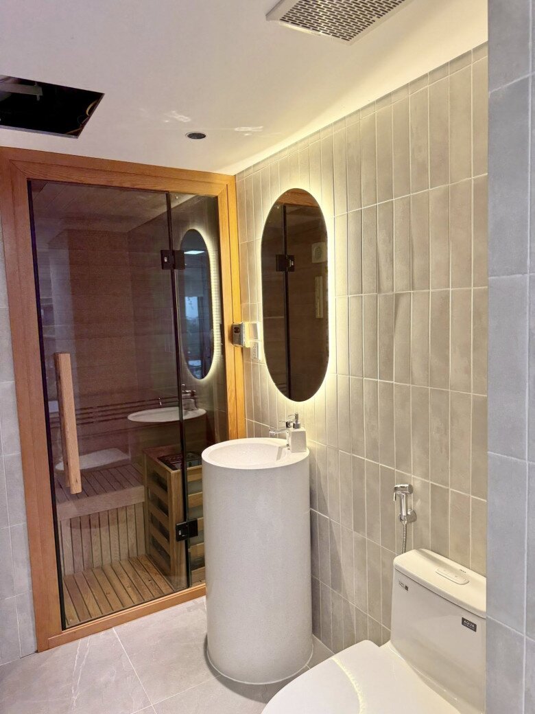 Thiết kế mang phong cách spa với phòng xông hơi gỗ, bồn rửa tròn lạ mắt và gương có đèn LED viền. Gạch ốp màu trung tính giúp không gian phòng tắm sáng và sạch sẽ.