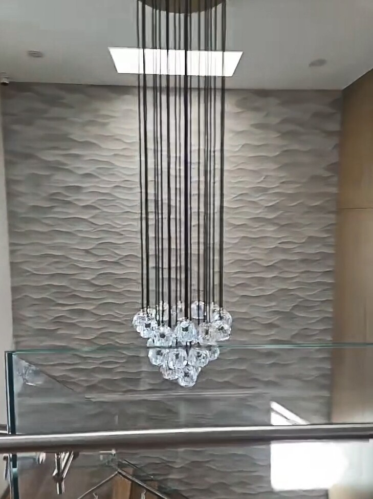 Đèn chùm pha lê treo giữa giếng trời, nổi bật trên nền tường 3D sóng lượn. Chi tiết này tạo cảm giác sang trọng cho toàn không gian, nhất là khi ánh sáng tự nhiên từ ô giếng trời phía trên chiếu xuyên qua pha lê.