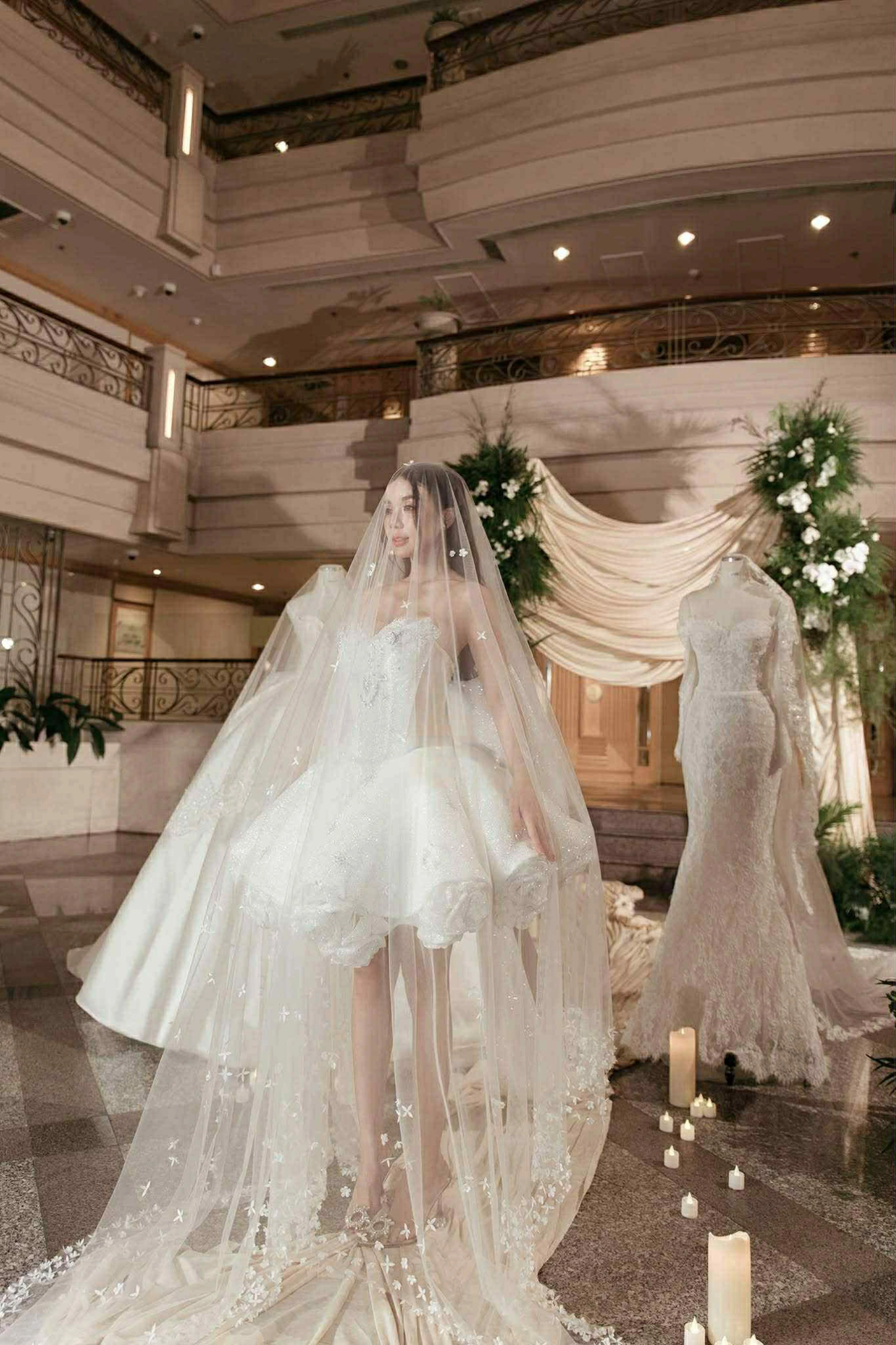 Điểm nhấn của tổng thể là lớp voan mỏng phủ ngoài dài chấm đất, thêu họa tiết hoa li ti dọc thân. Sự kết hợp giữa váy ngắn hiện đại và mạng che mặt truyền thống giúp cô dâu vẫn giữ được vẻ trang trọng cần có, nhưng không mất đi cá tính riêng.