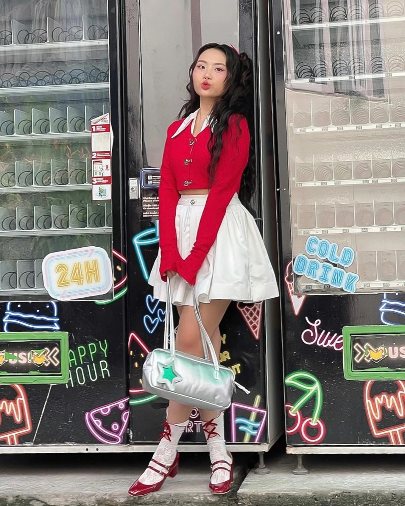 Với outfit gồm áo cardigan đỏ dáng lửng, chân váy trắng xếp ly, tất ren và giày Mary Jane đỏ, Phương Mỹ Chi hóa thân thành một “teen girl” đậm chất Hàn Quốc. Phụ kiện như túi xách ánh kim và tóc buộc nơ càng tăng thêm nét dễ thương nhưng không kém phần trendy. Sự hòa trộn giữa cổ điển và hiện đại tạo nên một diện mạo tươi mới, rất hợp với không khí đường phố sôi động.