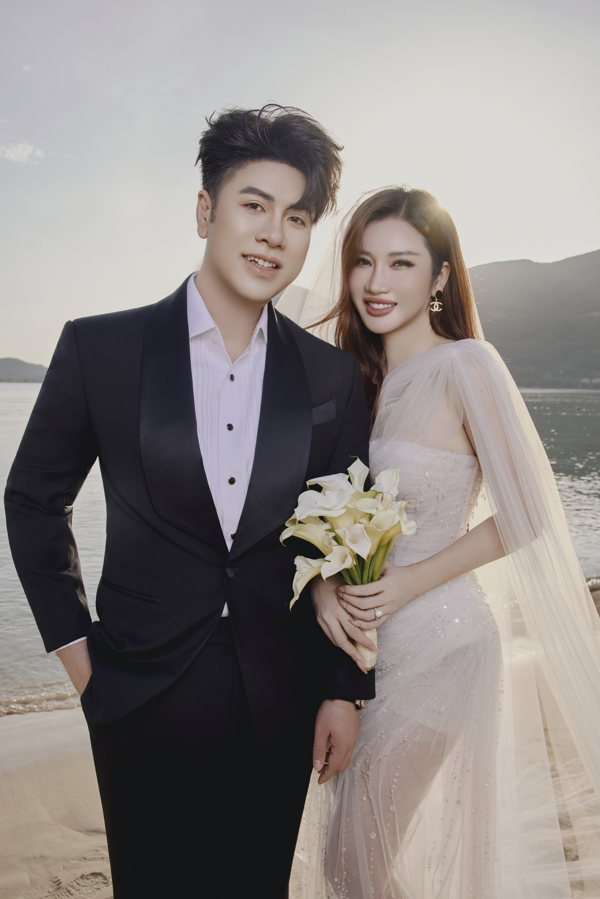 Trúc Phương diện hai thiết kế của Chung Thanh Phong trong buổi chụp. Trên bờ biển, cô chọn mẫu đầm ôm eo đính sequin và pha lê nhỏ li ti, phối cùng tay voan nhẹ và khăn voan dài - sự kết hợp giữa nét tinh khôi và chất lãng mạn.