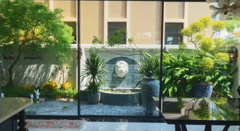 Góc nhìn từ bên trong nhà ra khoảng sân vườn phủ tràn màu xanh mát mắt qua hệ cửa kính lớn. Sự kết hợp giữa tiểu cảnh vòi nước chạm khắc đầu sư tử, thảm cỏ, lối đi lát đá và các chậu cây cảnh tạo nên một khung cảnh nghỉ dưỡng bình yên, thư thái ngay trong khuôn viên nhà.