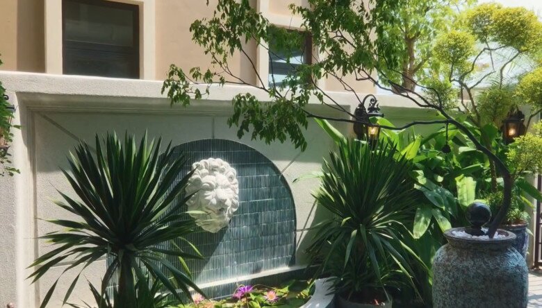 Sự kết hợp tinh tế giữa mảng tường đá chạm khắc nghệ thuật, vòi phun nước đầu sư tử và các loại cây cảnh tán rộng. Chiếc bình gốm lớn phong cách thô mộc đặt phía trước làm tăng thêm vẻ đẳng cấp, lịch lãm cho khuôn viên biệt thự.