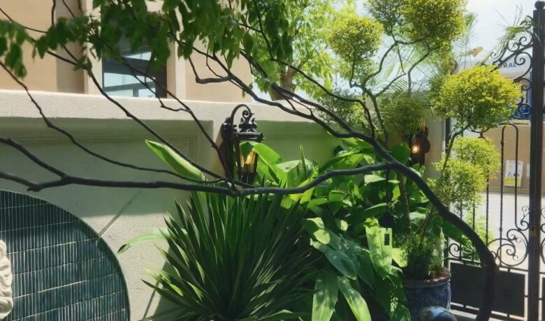 Góc nhìn cận cảnh từ phía cổng sắt uốn nghệ thuật hướng vào bên trong sân vườn biệt thự. Các tầng cây xanh được cắt tỉa gọn gàng, xen kẽ hệ thống đèn tường mang phong cách cổ điển, tạo vẻ đẹp lung linh và sang trọng ngay từ lối vào.