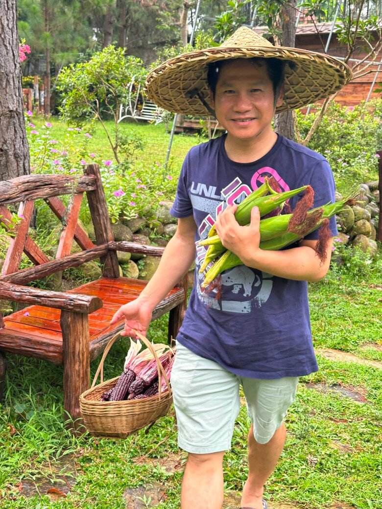 Một tay xách giỏ ngô tím, một tay ôm những bắp ngô tươi, nam ca sĩ hào hứng sải bước trên thảm cỏ xanh mướt rực rỡ sắc hoa ngoại ô. 