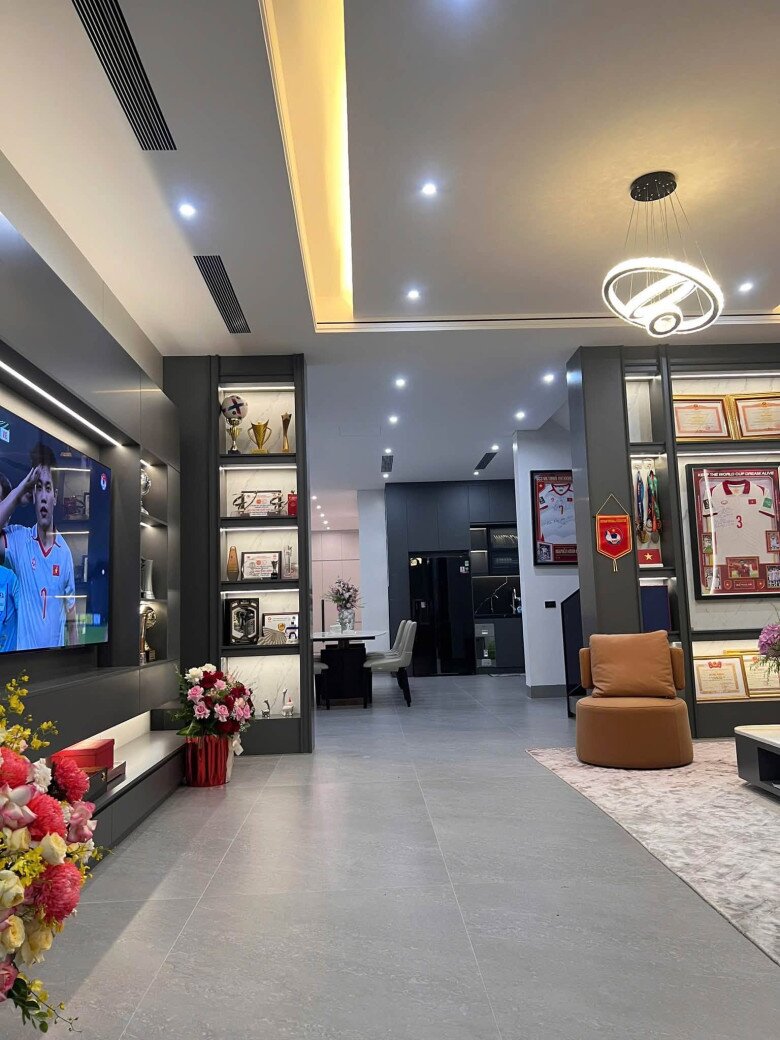 Phòng khách nổi bật với bộ sofa nỉ xám kem dáng thấp êm ái kết hợp cùng ghế đơn thư giãn màu cam đất tạo điểm nhấn màu sắc sinh động. Màn hình tivi cỡ lớn được đặt trung tâm hệ tủ ốp tường xám xước cao cấp, nơi gia đình thường quây quần theo dõi những trận cầu bùng nổ của nam cầu thủ.