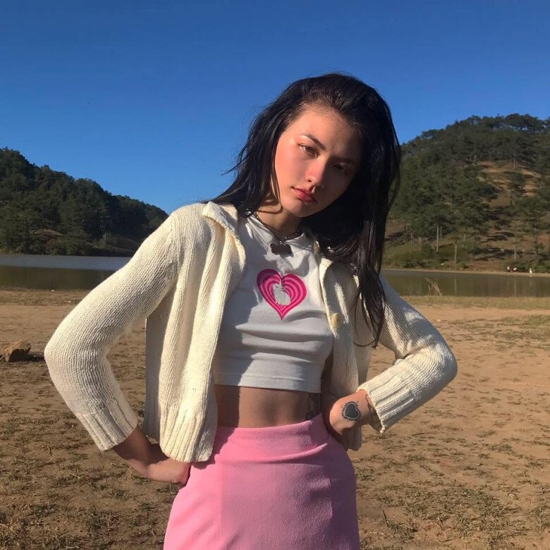 Trong khung hình này, Naomi chuyển sang gam hồng – trắng ngọt ngào với áo crop top thun gân ôm dáng, tạo điểm nhấn bằng họa tiết trái tim màu hồng neon nổi bật. Thiết kế ngắn giúp phần eo trở thành điểm nhấn tự nhiên, trong khi chân váy ôm cùng tông mang đến sự liền mạch cho set đồ.