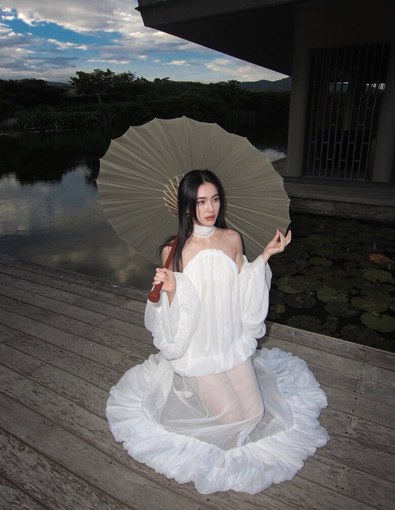 Một lần, “ma nữ đẹp nhất Thái Lan” chuyển sang phong cách nữ tính với thiết kế đầm trắng ngà từ chất liệu voan mỏng, dập ly bồng bềnh. Phần áo quây trễ vai tôn bờ vai và xương quai xanh, kết hợp dải lụa quấn cổ tạo điểm nhấn duyên dáng. Vạt váy xòe rộng phủ dài trên sàn cùng chiếc ô giấy truyền thống giúp Mai Davika mang đến vẻ đẹp vừa thanh thoát vừa cuốn hút.