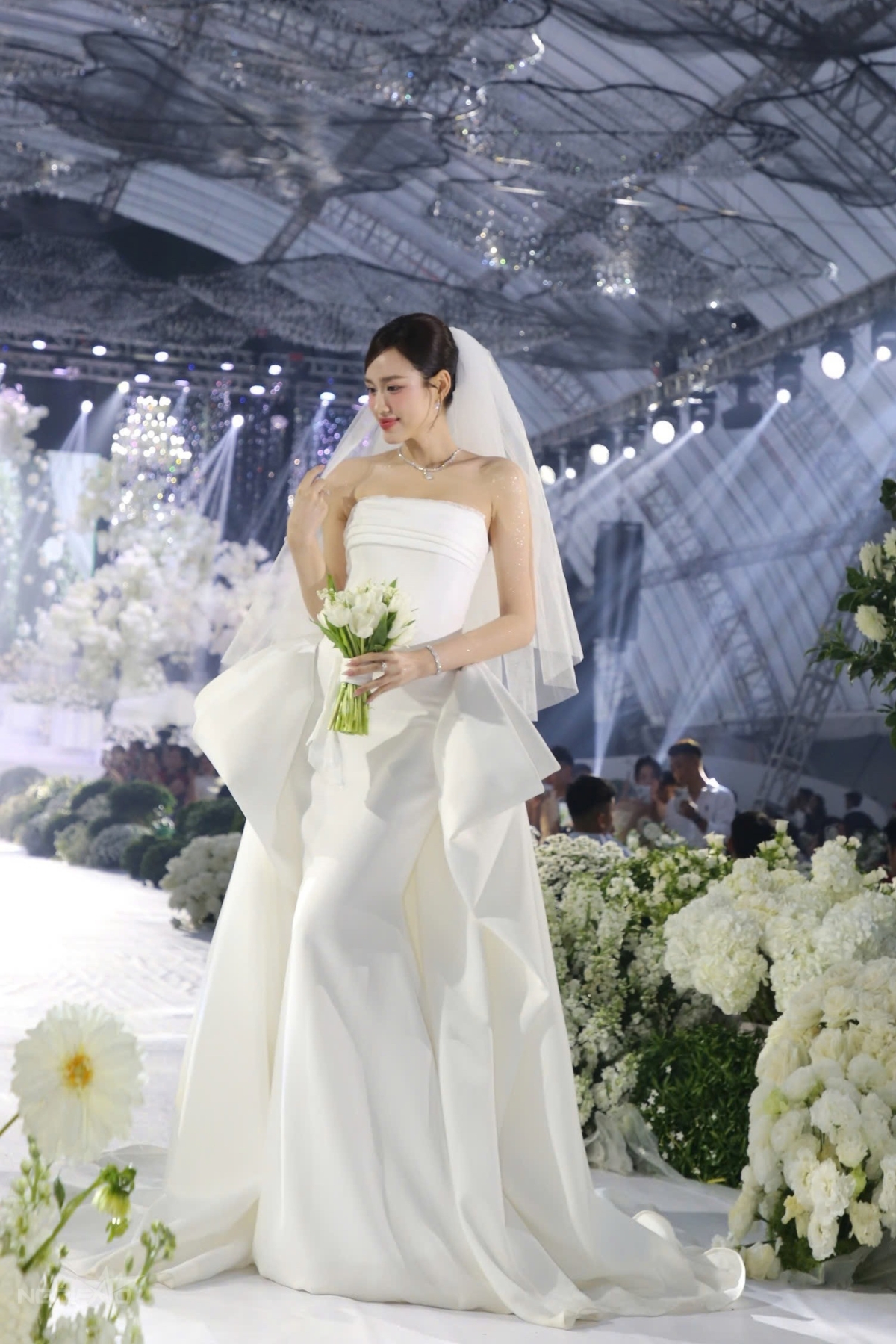 Đỗ Thị Hà chọn váy của NTK Lê Thanh Hòa để chào đón quan khách đến với tiệc cưới. Thiết kế được thực hiện từ chất liệu mikado cao cấp, không sử dụng bất kỳ chi tiết đính kết nào mà chỉ dựa vào kỹ thuật dựng phom và xử lý bề mặt vải để thể hiện sự sang trọng, thuần khiết. Ảnh: Đắc Thành