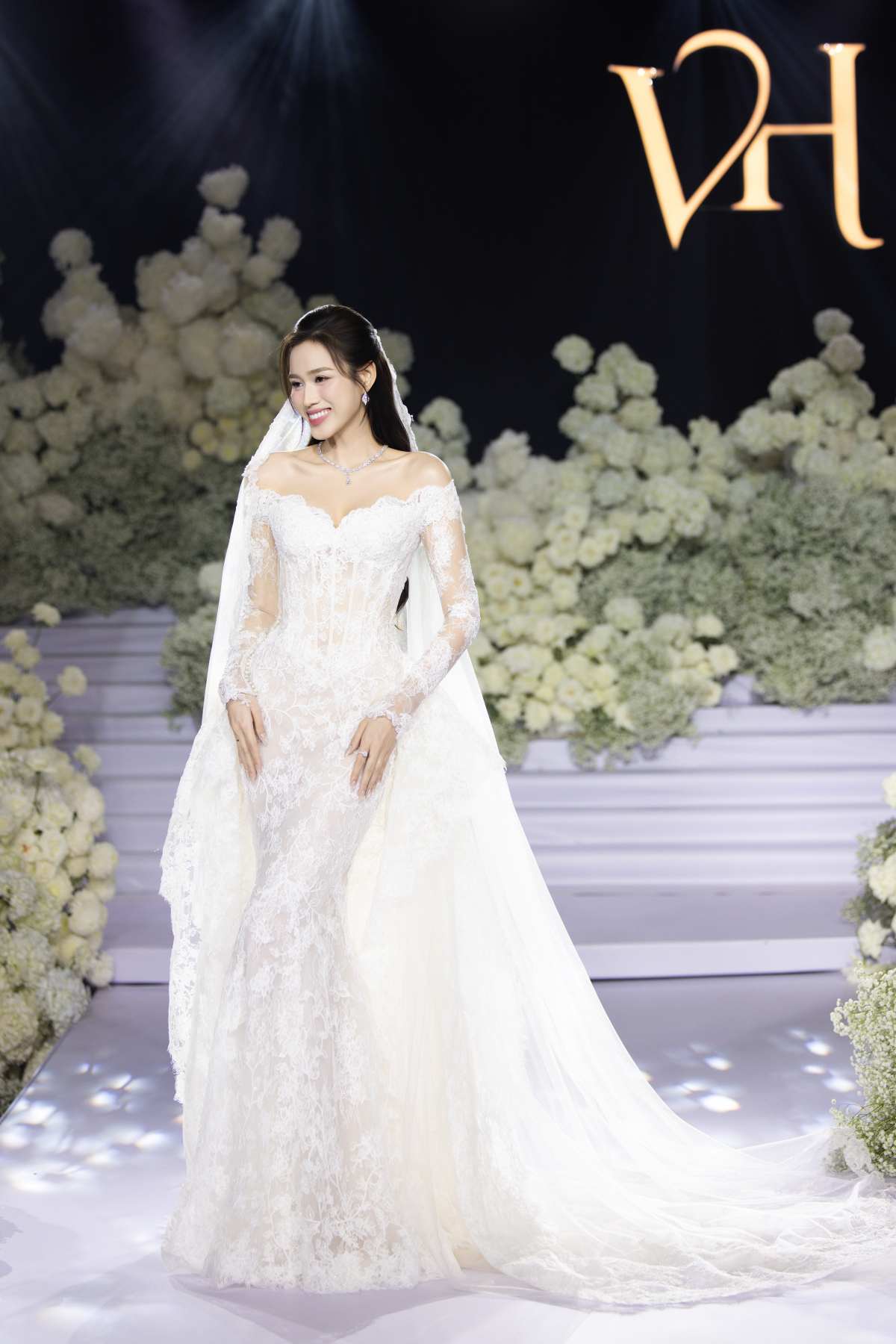 Thiết kế tạo nên từ ren tơ Pháp cao cấp, ôm trọn đường cong của Đỗ Thị Hà. Phần eo tạo phom corset siết chặt vòng hai nhưng vẫn giữ được sự mềm mại. Đầm đi kèm khăn voan dài cả chục mét, phù hợp không gian cưới cổ tích.