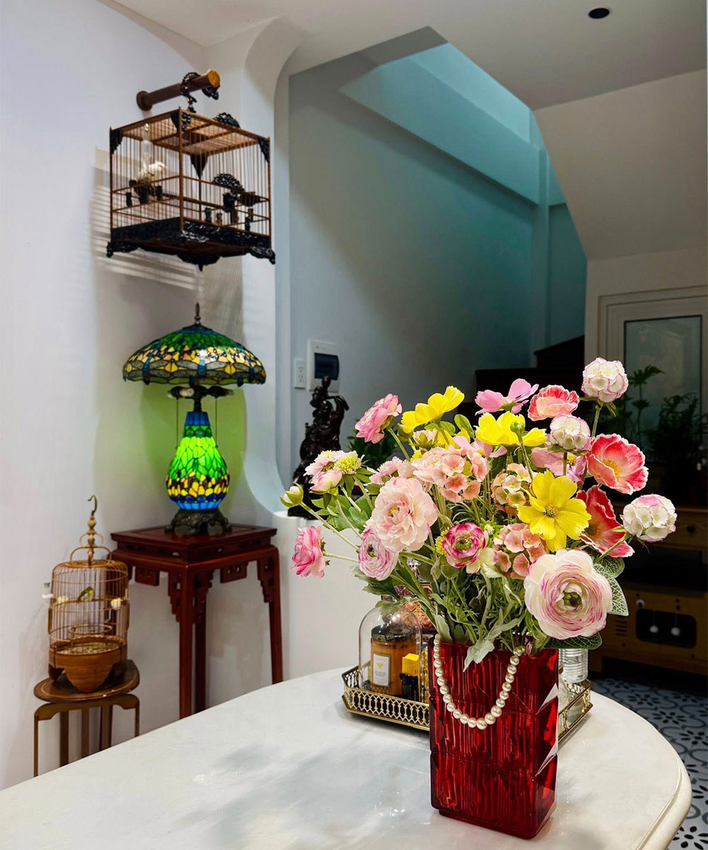 Phòng khách nhỏ nhưng ấm cúng, với bàn trà bầu dục, đèn Tiffany màu sắc cổ điển, lồng chim treo cạnh cửa sổ và nhiều tranh hội họa tươi sáng.