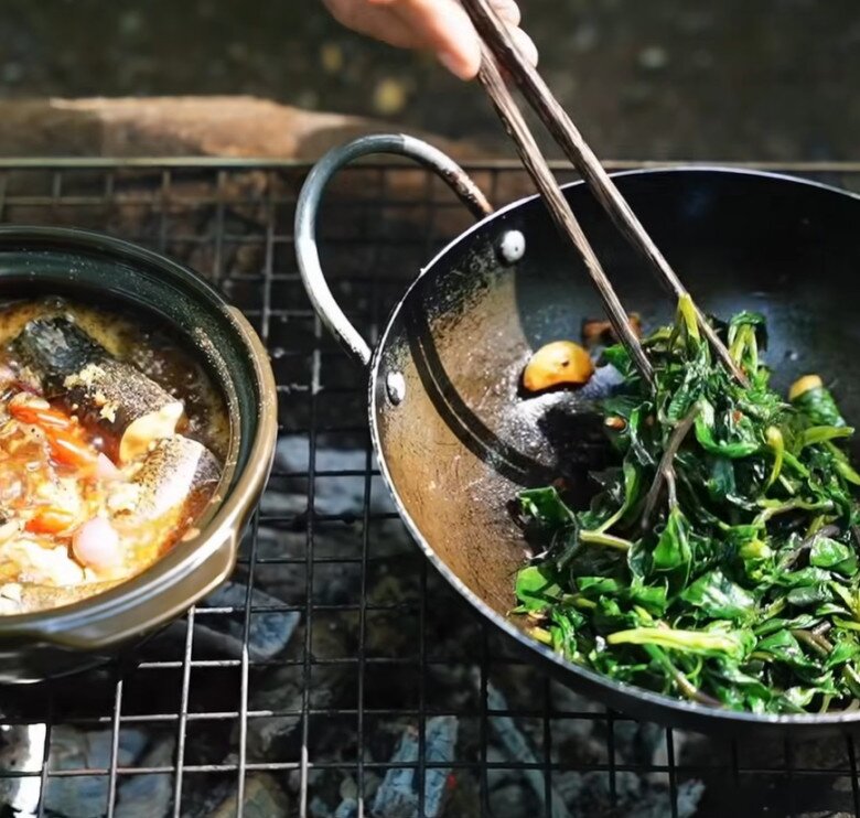 Hai món ăn đang được nấu song song trên bếp lửa - cá kho đậm đà và rau xào tỏi thơm phức, tạo nên hương vị đồng quê mộc mạc nhưng tròn vị.