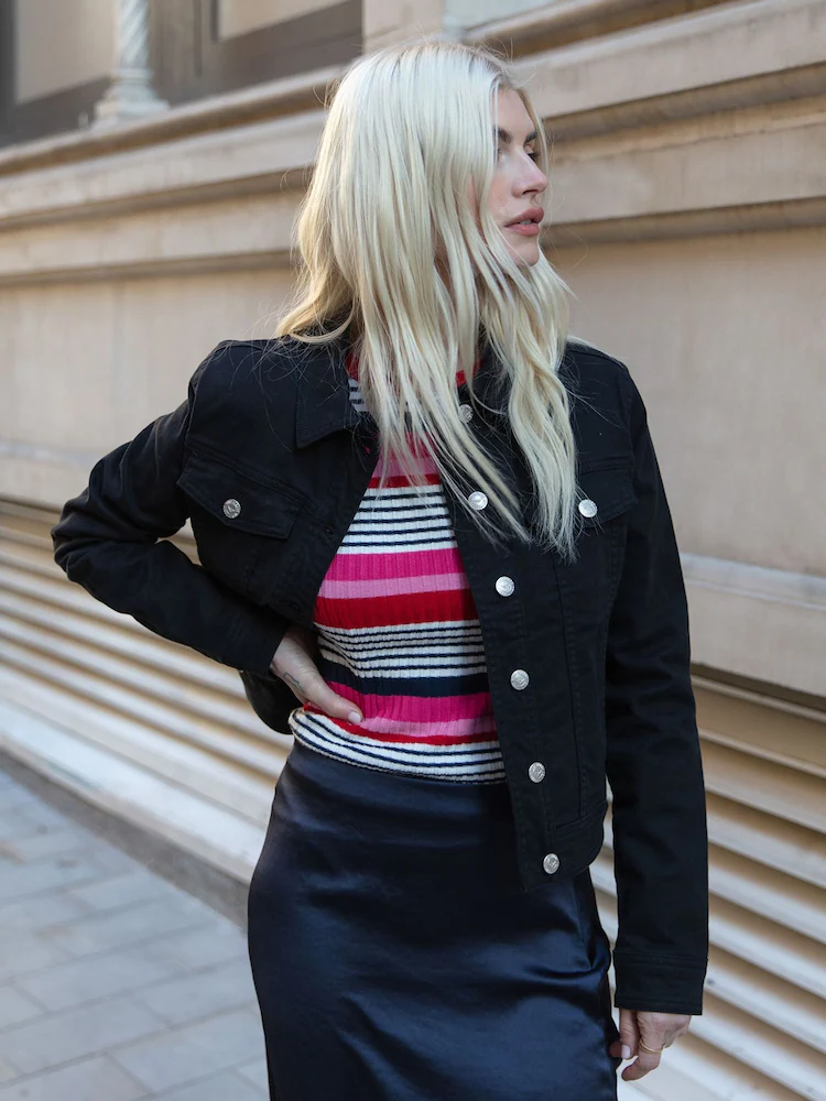 Nổi bật là áo khoác đen dáng jacket với phom cứng cáp, mạnh mẽ. Khi kết hợp cùng áo len kẻ nhiều màu và chân váy tối màu, set đồ tạo nên sự tương phản thú vị, vừa cá tính vừa thời thượng.