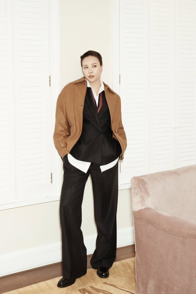 Đàm Thu Trang khoác thêm áo jacket nâu bên ngoài bộ suit tối màu, tạo chiều sâu và sự đối lập thú vị về sắc độ. Cô đứng thẳng, tay đút túi, mang đến vibe tổng tài đầy chủ động và tự tin.