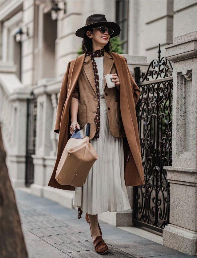 Áo khoác dạ camel dáng dài được khoác hờ trên vai, bên trong là áo blazer tông nâu nhạt kết hợp cùng chân váy midi xếp ly màu trắng sữa, mang đến sự tương phản dịu mắt giữa chất liệu dày ấm và bề mặt mềm mại, bay nhẹ của váy.