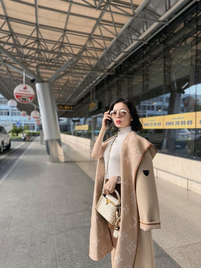 Tại sân bay, Lệ Quyên hóa thân thành quý cô thành thị thanh lịch trong chiếc áo khoác Louis Vuitton tông beige trứ danh, phủ kín logo monogram cổ điển. Cô kết hợp khéo léo với áo cổ lọ màu kem và túi xách da cá sấu sáng màu, tạo nên tổng thể tiệp sắc tinh tế.