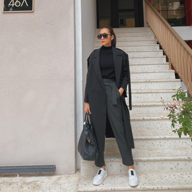 Một set đồ đen toàn tập nhưng không hề nhàm chán: áo cổ lọ ôm nhẹ mix cùng quần cạp cao và trench coat đen dáng dài đầy quyền lực. Giày slip-on trắng tạo điểm sáng tinh tế, trong khi túi xách size lớn tăng tính thực dụng mà vẫn thanh lịch. Đây là minh chứng cho khả năng tối giản tuyệt vời của Hà Tăng.