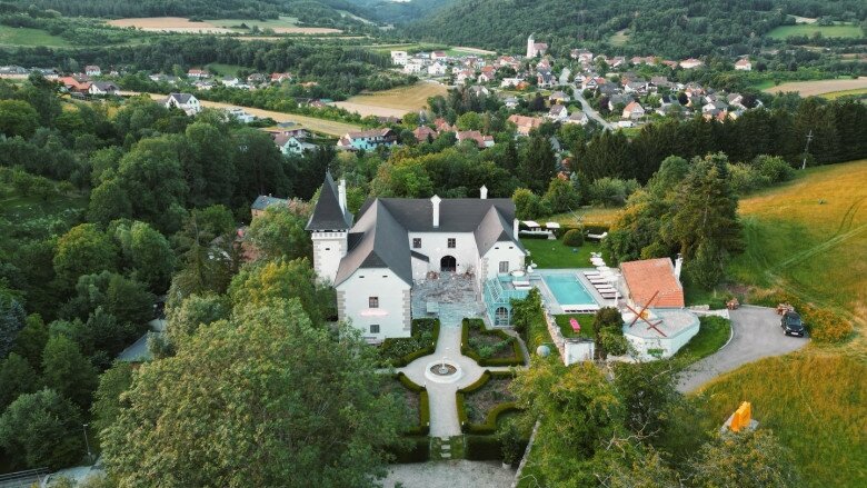 Toàn cảnh biệt thự từ trên cao. Công trình có cấu trúc như lâu đài châu Âu cổ điển với hai tháp nhọn, khuôn viên xanh rộng, nằm giữa một thung lũng yên bình tại vùng quê nước Áo. Hồ bơi nằm sát nhà chính, bao quanh bởi rừng cây và đồng cỏ.