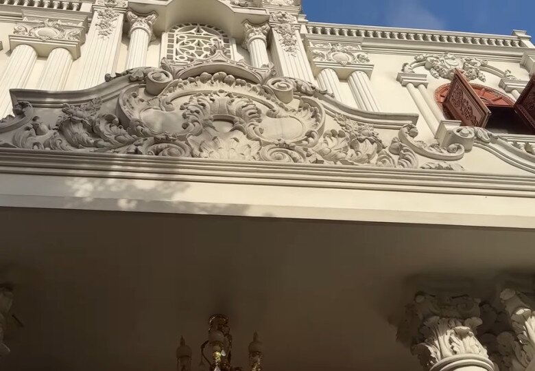 Sảnh chính lộng lẫy với trần cao, hoa văn đắp nổi và tường sơn vàng kem, tạo cảm giác như bước vào một lâu đài cổ. Mặt tiền tầng trên của biệt thự, thể hiện sự cầu kỳ trong từng chi tiết phào chỉ và phù điêu trang trí.