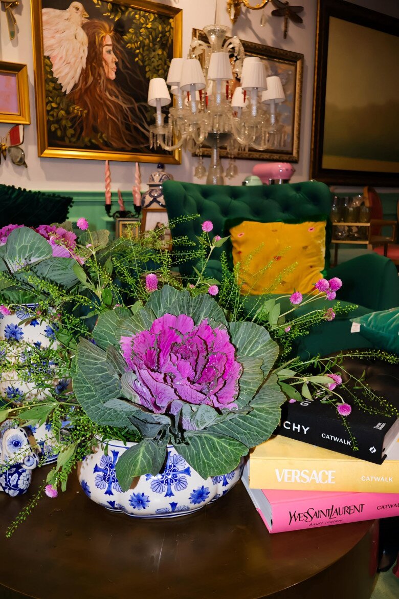 Góc phòng nổi bật với hai lọ hoa bắp cải đặt cạnh nhau, hài hoà với nội thất như ghế nhung xanh rêu, gối vàng thêu tay và tường xanh pastel, tạo nên khung cảnh như một bức tranh sống động.