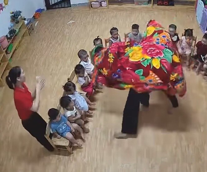 2 cô giáo dùng chậu làm đầu sư tử, quàng chăn lên người để nhảy múa, 1 cô giáo khác đứng bên cạnh cổ vũ.